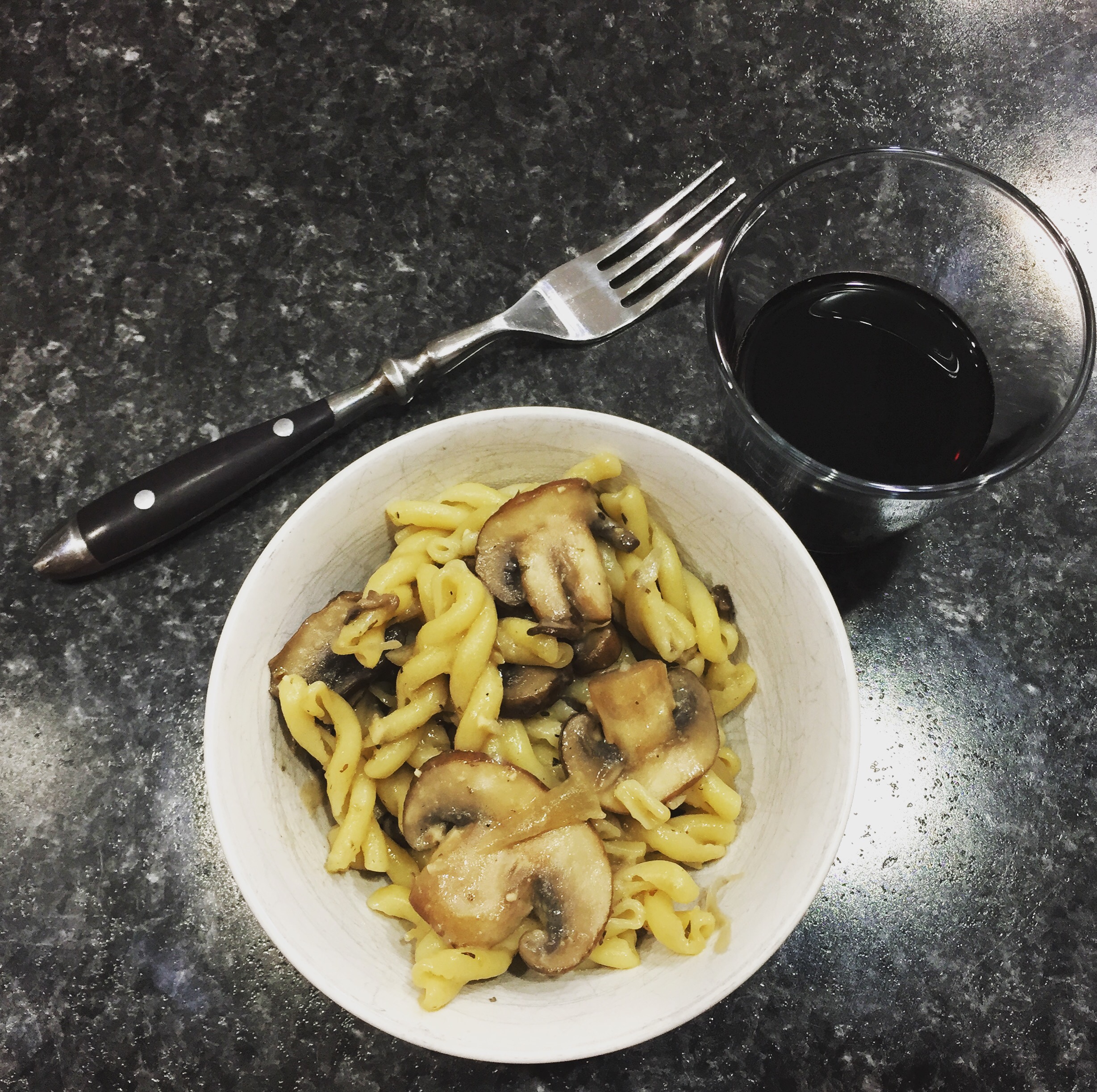 A top-down image of a white bowl filled with pasta and mushrooms, with a fork sitting next to it and a glass of red wine.
