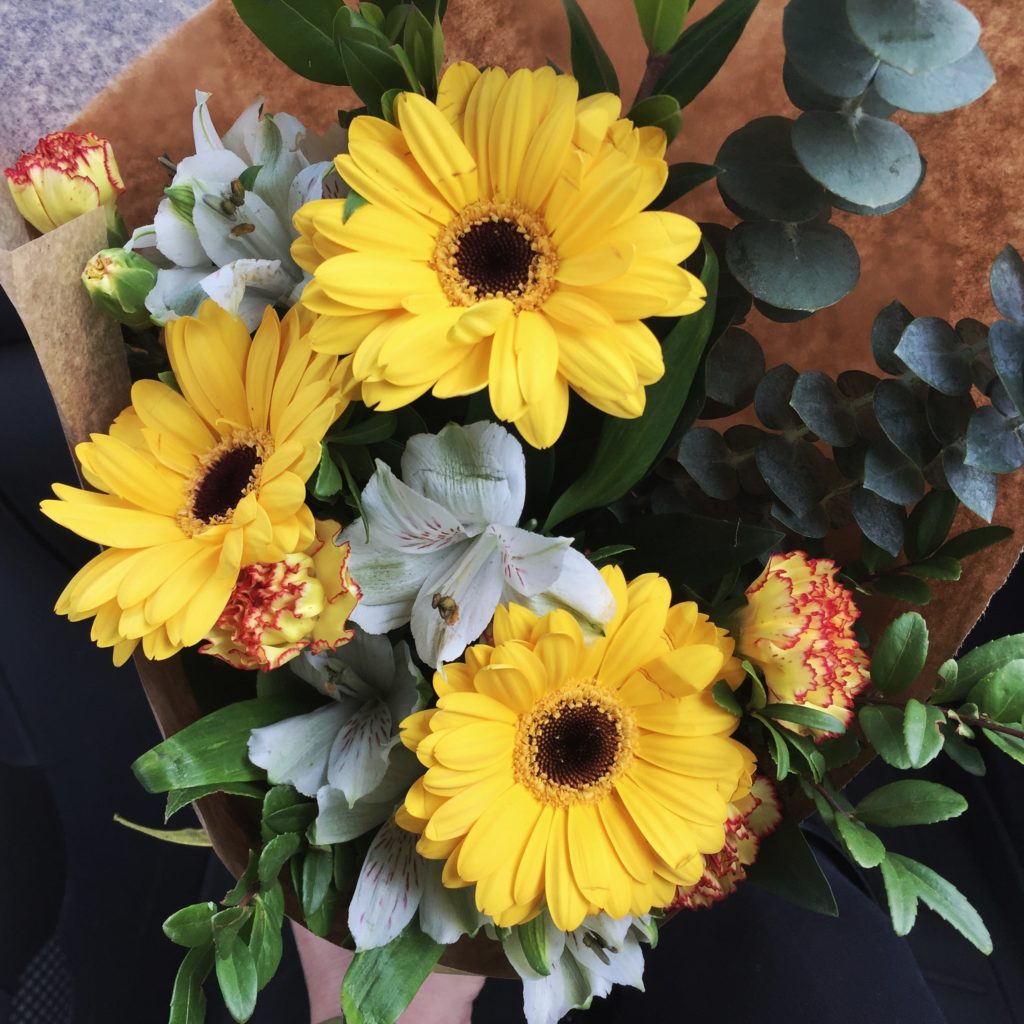 A bouquet of gerbera daisies with some carnations and mixed green plant material, wrapped in paper with a yellow bow.