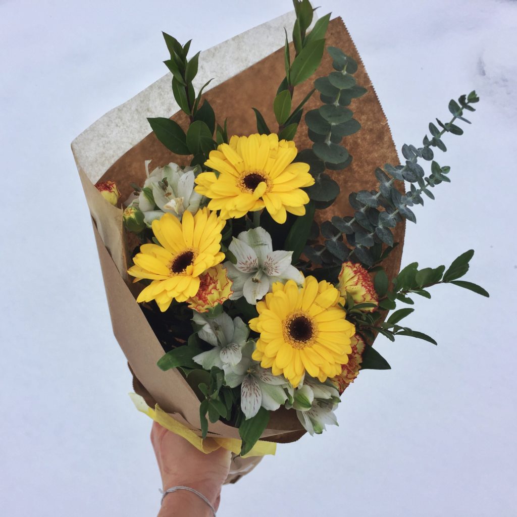A bouquet of gerbera daisies with mixed plant material wrapped in paper with no plastic, against a snowy backdrop in winter.