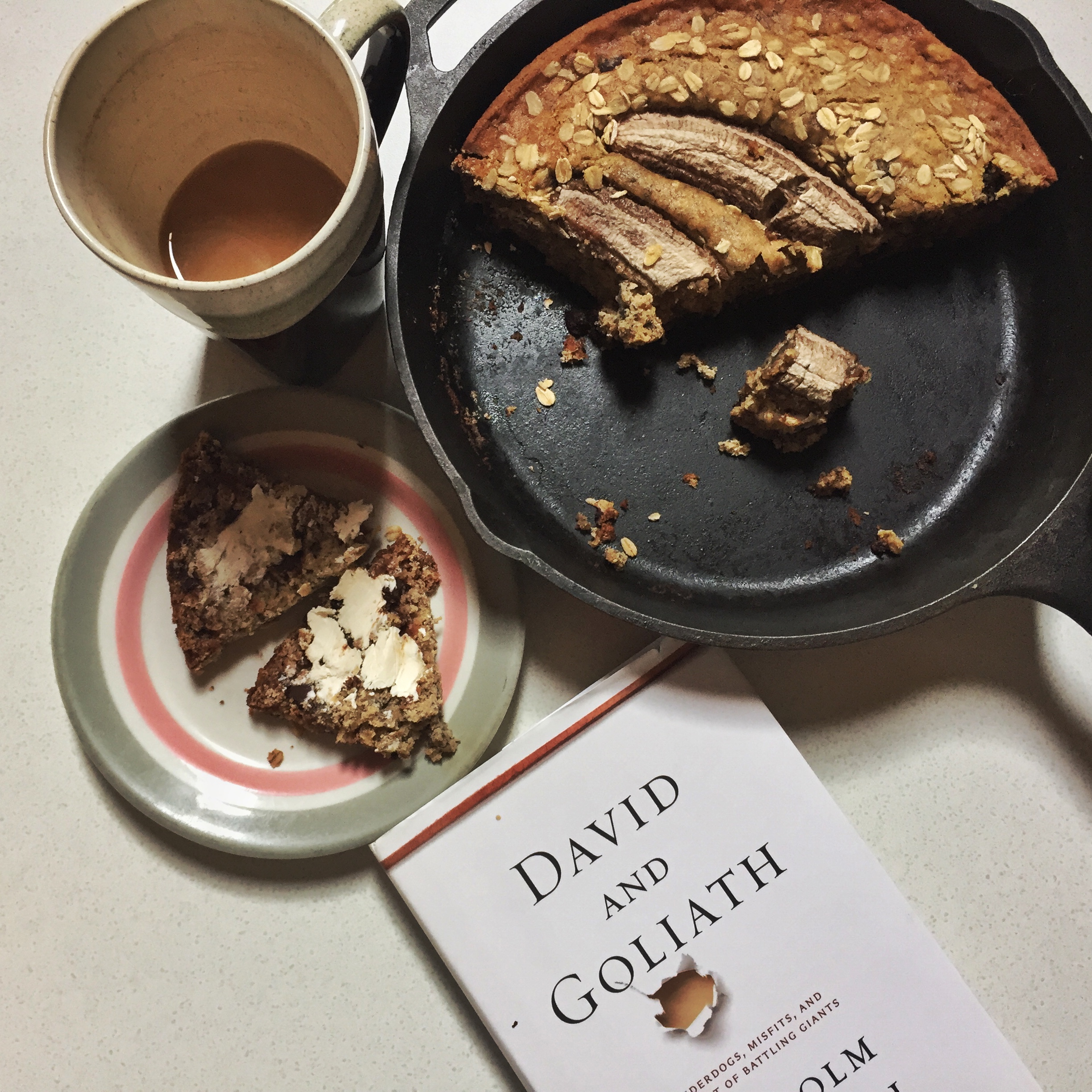 flat lay photo of a cast iron pan full of banana bread alongside a mug of coffee and the book David and Goliath by Malcolm Gladwell