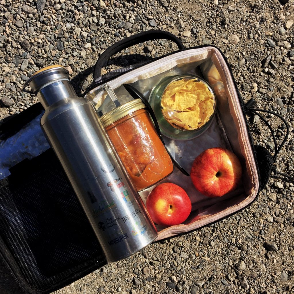 a visual depiction of small acts leading to big change: a lunchbox filled with a jar of soup, a jar of crackers, fruit, and a reusable water bottle. Zero waste lunch.