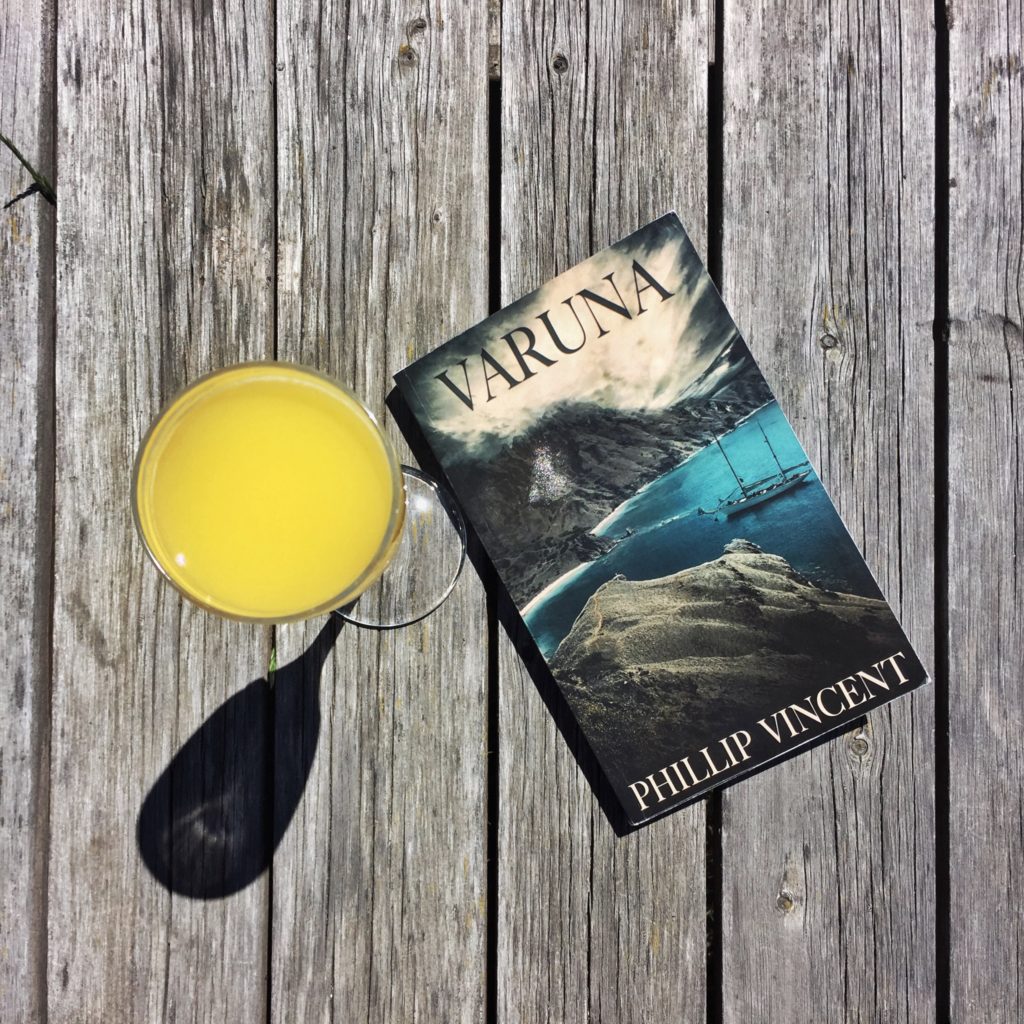 A copy of Varuna, the debut novel by author Phillip Vincent, sitting on a wooden bench with a champagne flute of orange juice next to it.