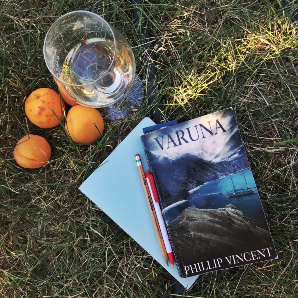 A copy of Varuna, the debut novel from author Phillip Vincent, laid out on the grass with a notebook, pen, glass of wine, and a few apricots.