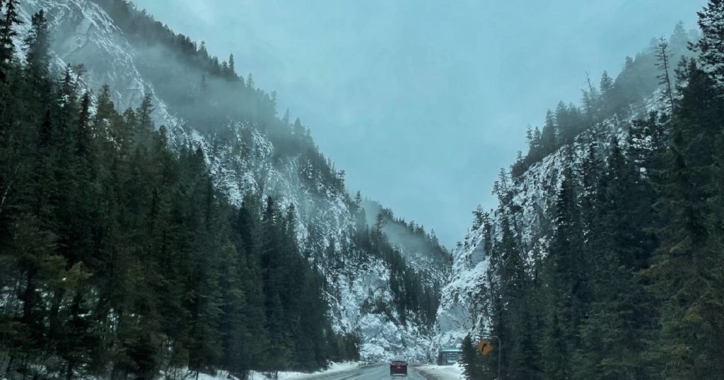 Slow and steady through wintery mountain passes, this image depicts a highway cutting through a valley surrounded by rocky mountains and pine tree forests on a foggy day.