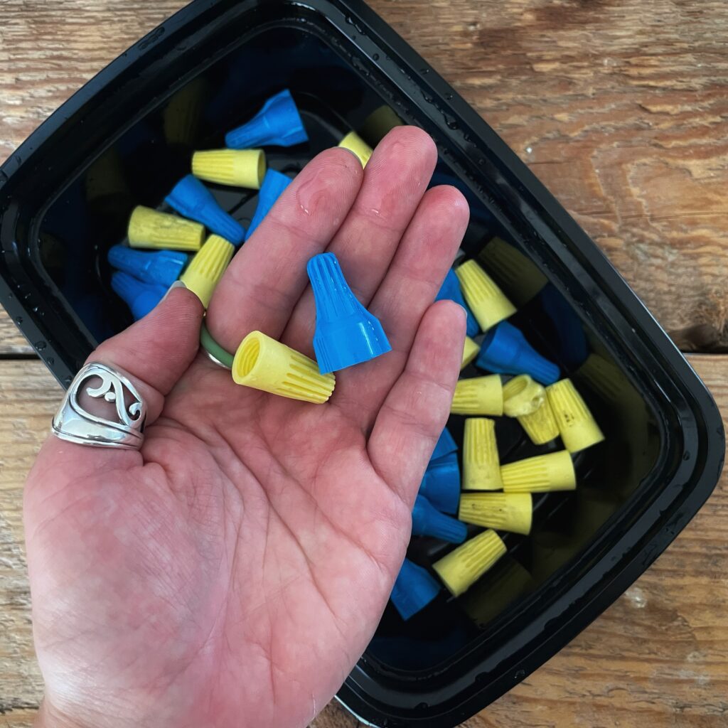 Author's hand holding two plastic twist-on wire connectors in blue and yellow colours, over a black plastic container filled with similar material.