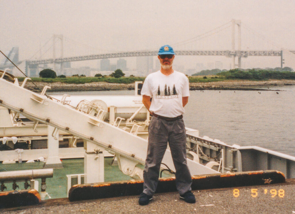 John Williams stands in front of a research vessel in Japan, with his arms behind his back, wearing a white shirt, blue ball cap, and grey pants.