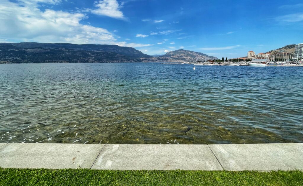 The view of Okanagan Lake from Kelowna's City Park, with sidewalk and grass in the foreground and the downtown waterfront and West Kelowna hillside in the background.