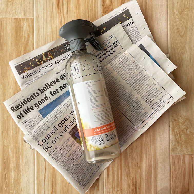 A reused plastic spray bottle filled with DIY glass cleaner lays on top of a pile of newspapers.