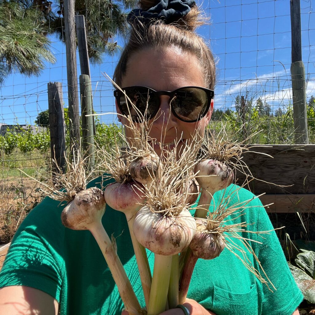A selfie of the author wearing a green shirt and black sunglasses, holding bunches of garlic bulbs in front of her face.