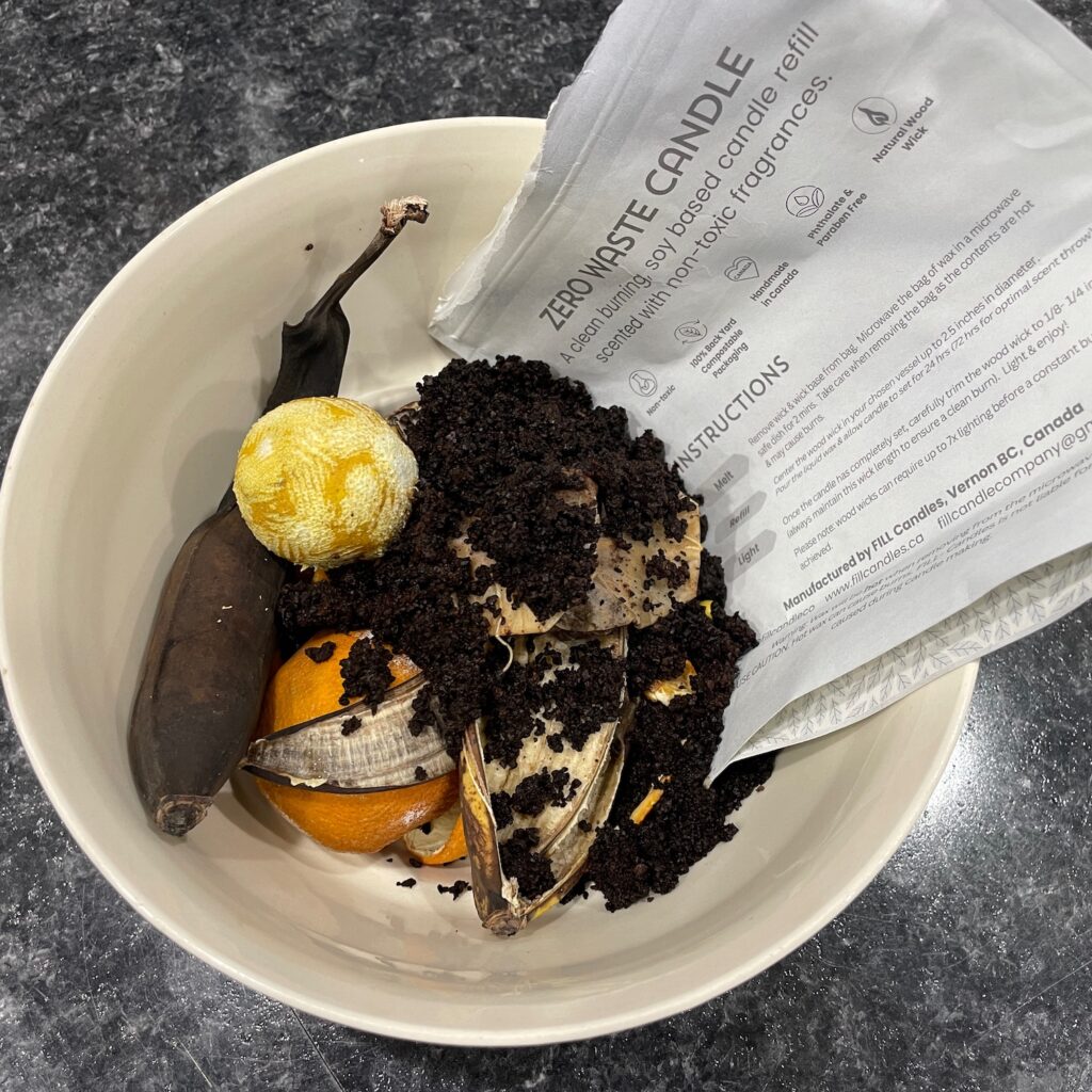 A bowl of items to be composted including a banana, coffee grounds, orange peels, and the bag from a candle refill kit.