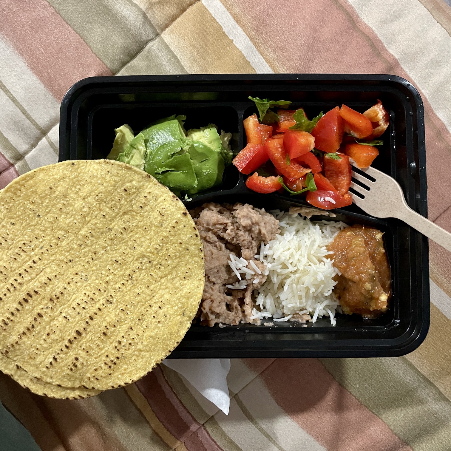 A black plastic takeout container is filled with fresh avocado, red bell pepper salad, rice, beans, and salsa along with yellow corn tostadas.