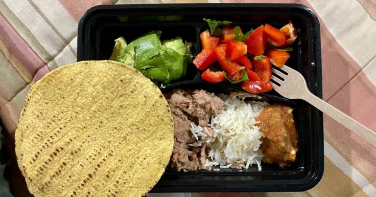A black plastic takeout container is filled with fresh avocado, red bell pepper salad, rice, beans, and salsa along with yellow corn tostadas.