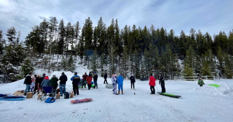 A group of people stand at the bottom of a forested hillside wearing winter clothes, with sleds scattered around them.