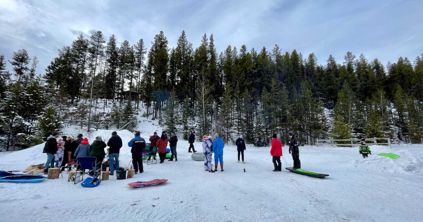 A group of people stand at the bottom of a forested hillside wearing winter clothes, with sleds scattered around them.