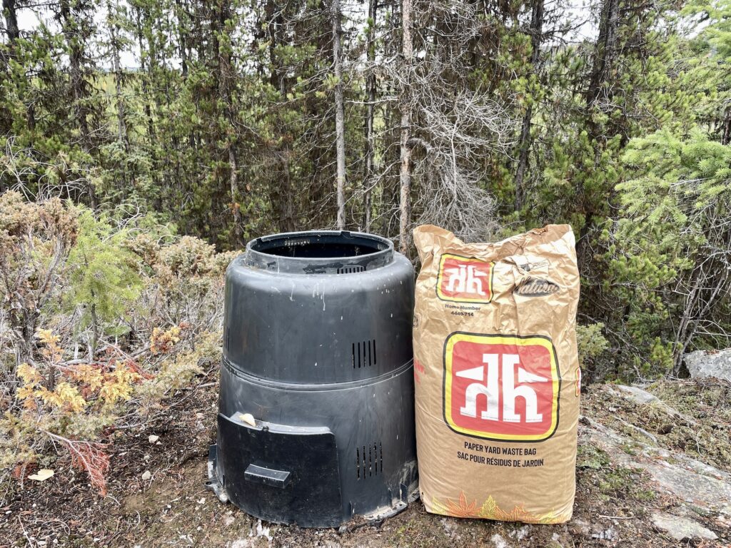 A black home composter sits next to a paper leaf bag from Home Hardware, in the forest.
