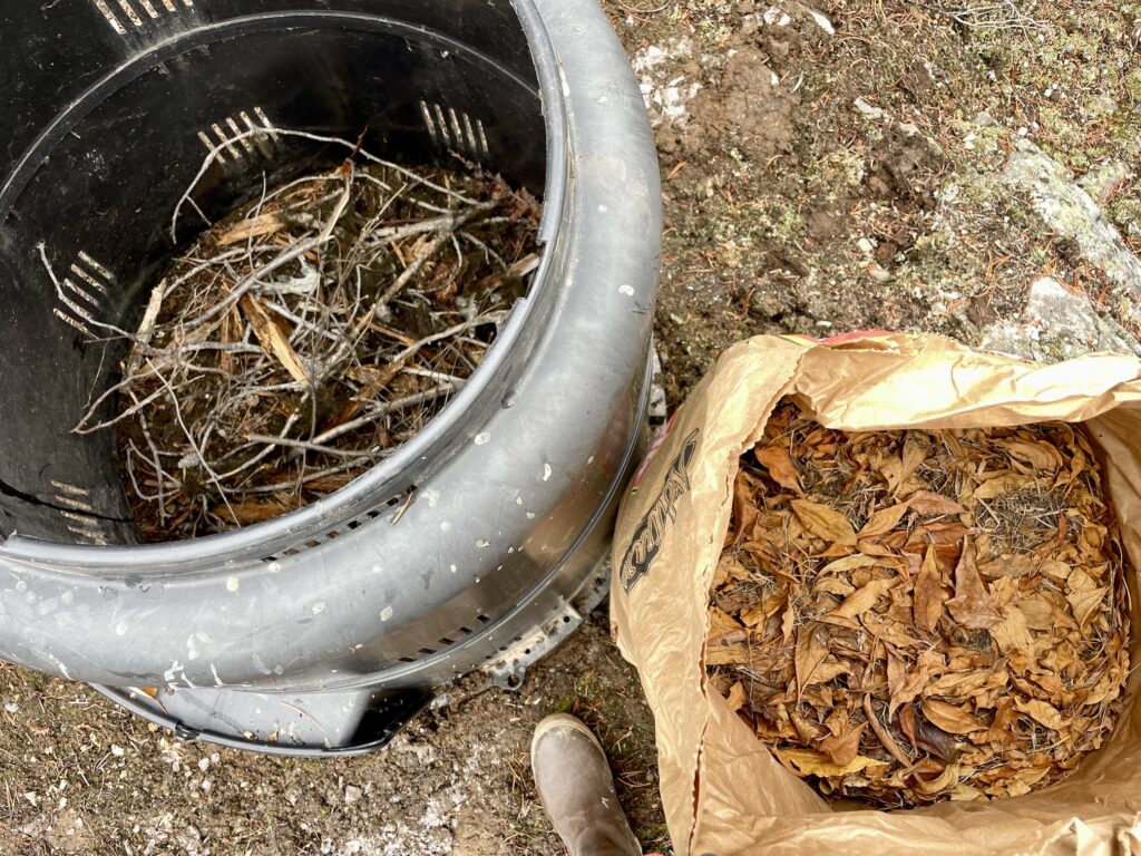 A black plastic home composter is shown from the top down, with the composter on the left - containing a pile of twigs - and a bag of leaves on the right.