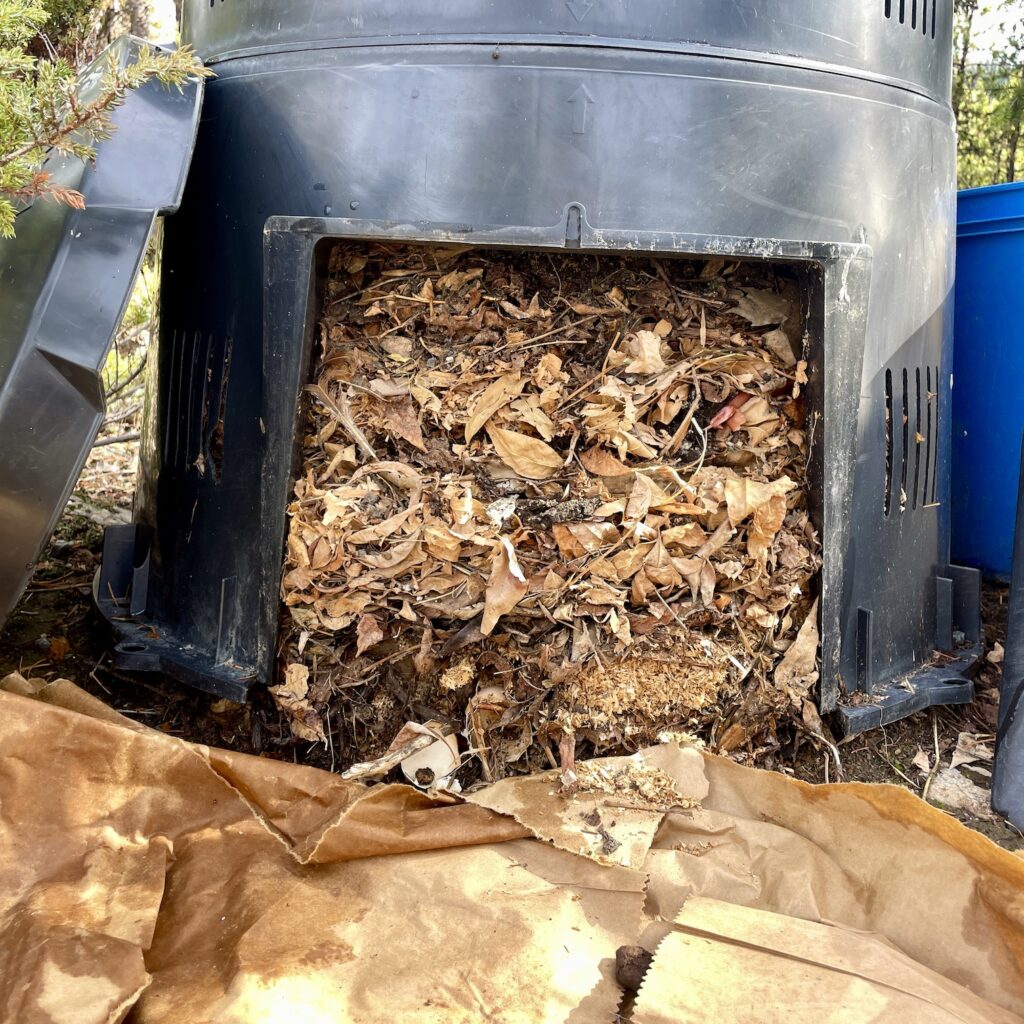 A black composter is shown with a side panel removed, revealing compost inside.
