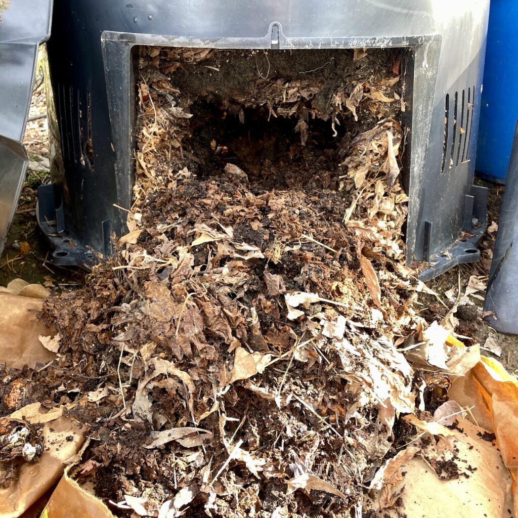 A black composter is shown with a side panel removed, revealing compost inside.